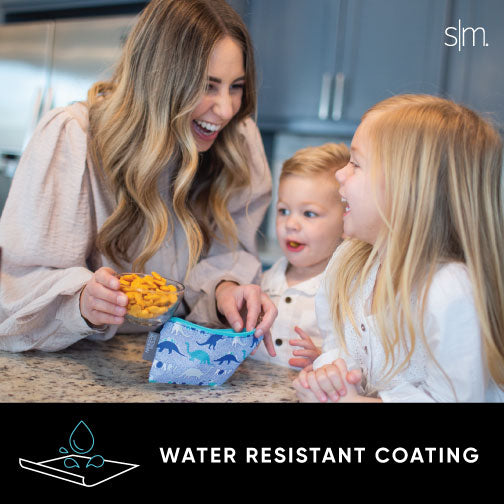 Woman in beige cardigan with two children in white shirts in a light gray kitchen, holding Ellie Snack Bags featuring warm neutral tones and bright color accents.