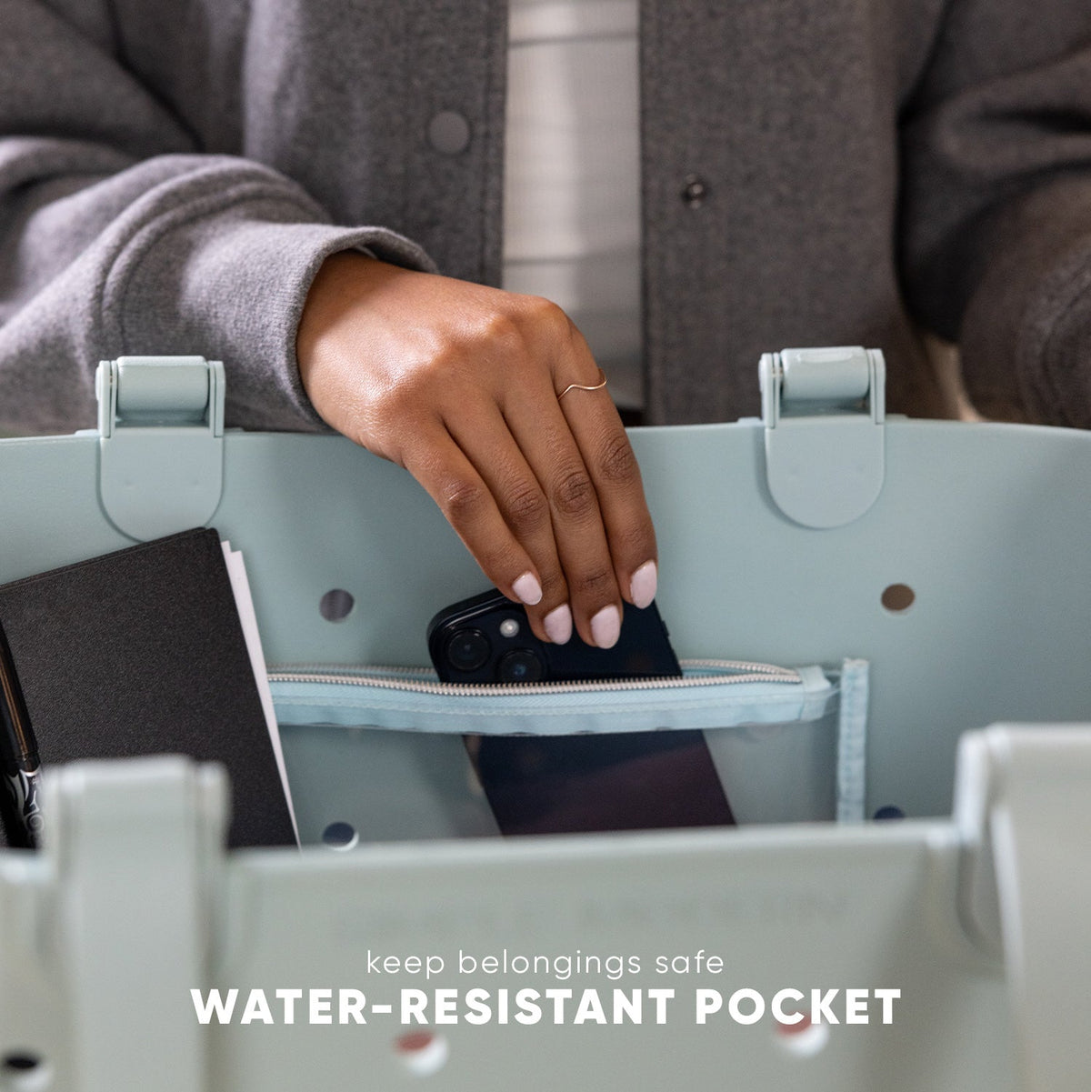 Person placing black smartphone into sage green tote bag's water-resistant pocket. Text reads "keep belongings safe WATER-RESISTANT POCKET." Lifestyle product demonstration showing bag functionality.
